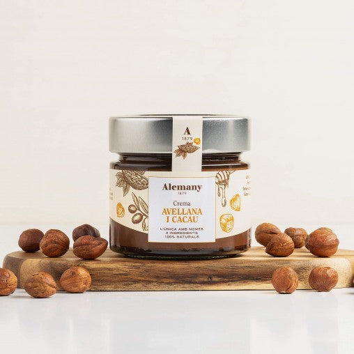 Alemany Hasselnøddecreme med honning og kakao i glas, på et brædt med hasselnødder omkring.